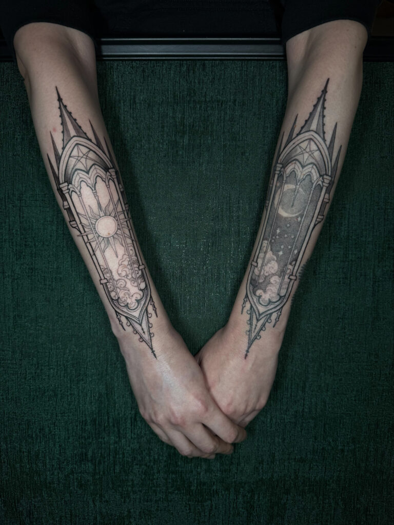 Symmetrische tattoos op beide onderarmen in de vorm van neogotische kerkvensters. Het linker venster toont een stralende zon met wolken, het rechter venster een halve maan met een sterrenhemel en wolken. Een prachtig staaltje fineline blackwork van Maeve (Maeve Sanaa) op de Sint-Niklaas Tattoo Expo.