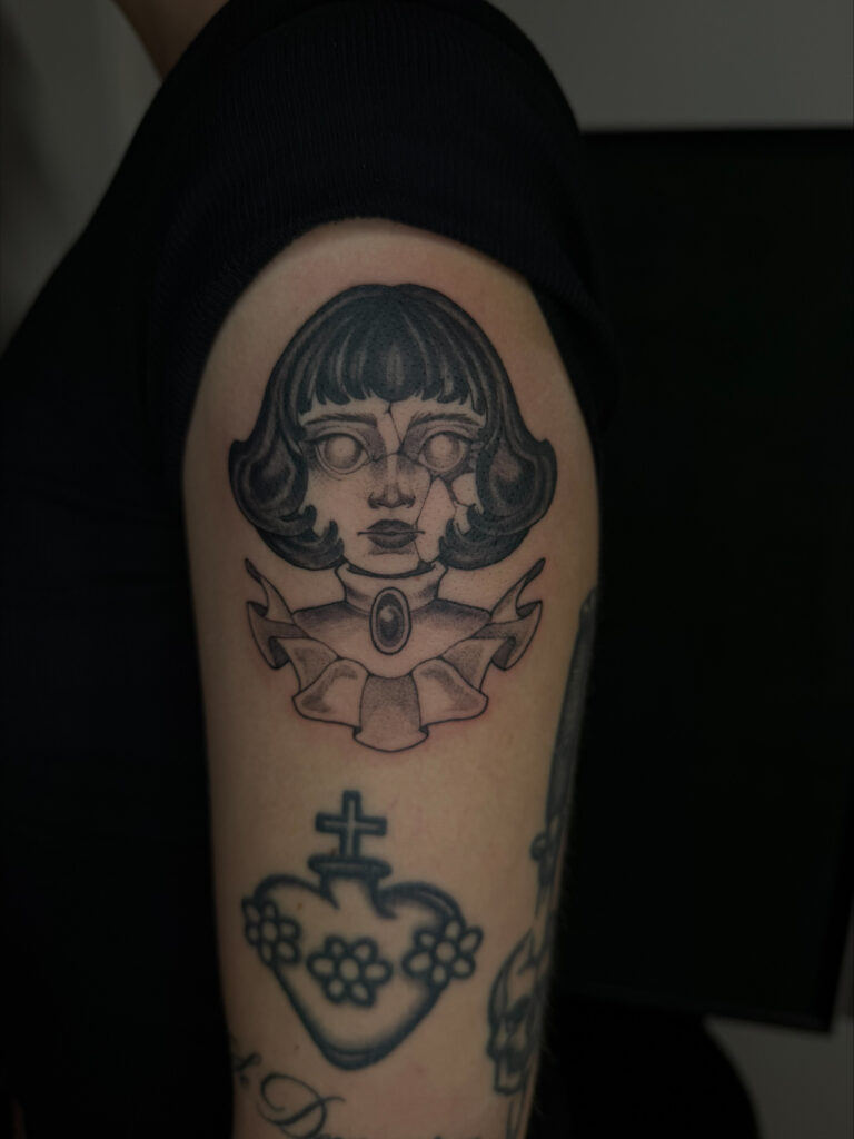 Een karakteristieke black and grey tattoo op een bovenarm van een porseleinen poppenhoofd met barsten en witte ogen zonder pupillen. De pop draagt een sierlijke kraag met een edelsteen. Werk van Maeve (Maeve Sanaa) tijdens de Sint-Niklaas Tattoo Expo.