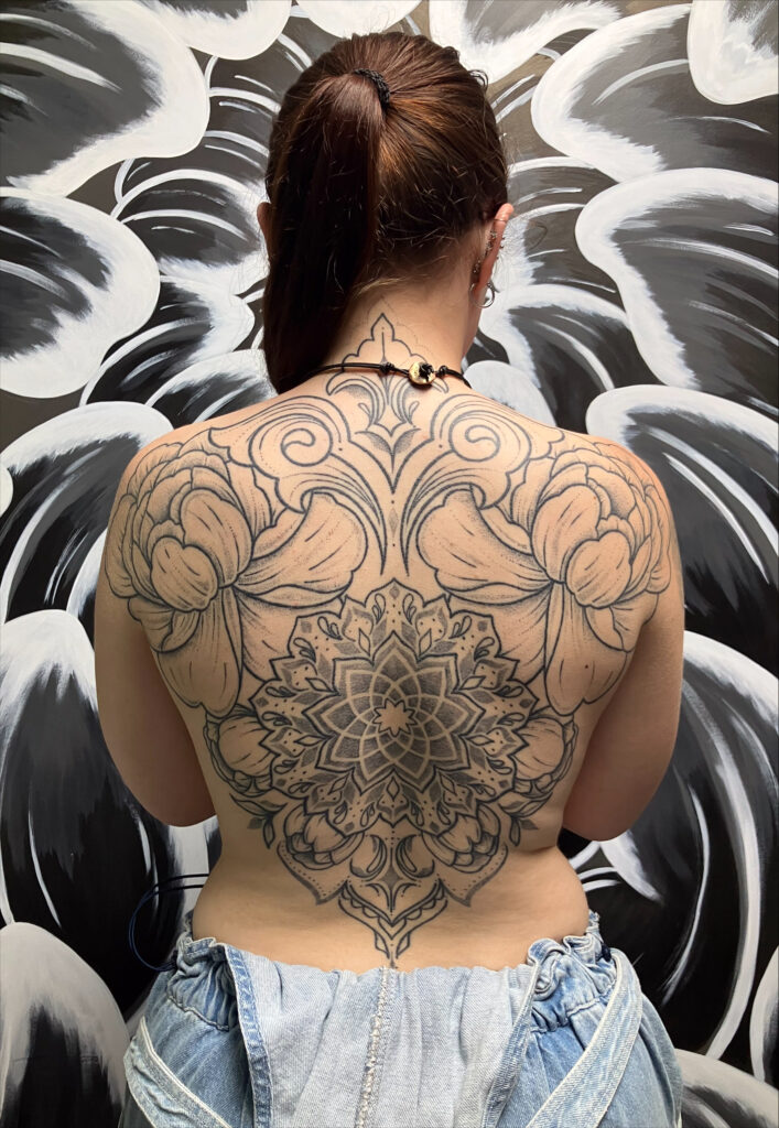 Een indrukwekkend groot rugstuk bestaande uit een centrale, gedetailleerde mandala onderaan, die overgaat in symmetrische florale en krullende ornamenten over de schouderbladen en tot in de nek. Een meesterwerk in ornamenteel blackwork door Monica van Poked by Monique op de Sint-Niklaas Tattoo Expo.
