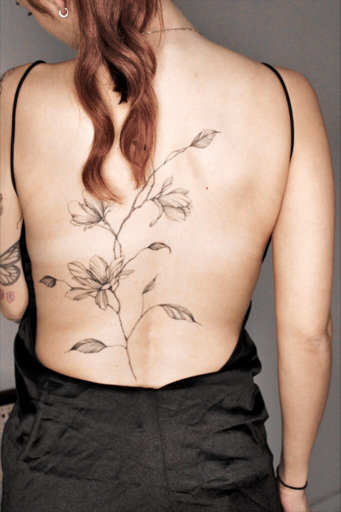 Een verfijnde fineline tattoo van een elegante bloementak die verticaal over de rug loopt. Het ontwerp is subtiel en volgt de natuurlijke lijnen van het lichaam, uitgevoerd met uiterst dunne lijnen en zacht schaduwwerk. Werk van Marina (Serial Chillers) op de Sint-Niklaas Tattoo Expo.