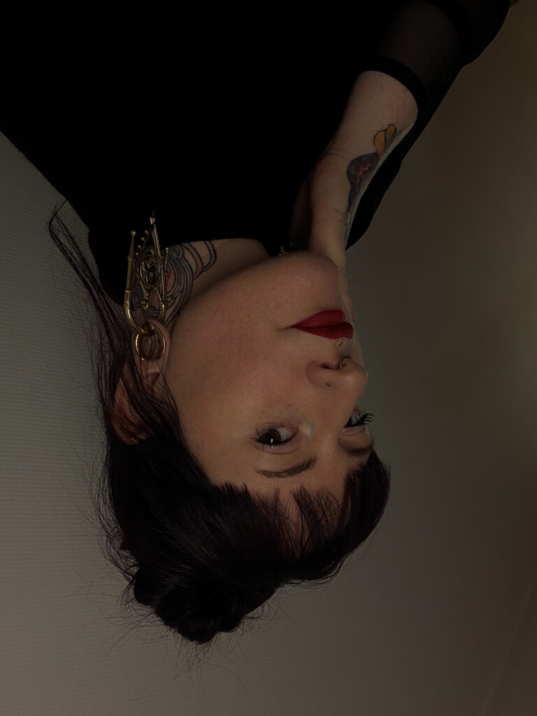 Een artistiek portret van tattoo-artiest Maeve (Maeve Sanaa) op de Sint-Niklaas Tattoo Expo. Ze heeft donker haar met een rechte pony, draagt opvallende goudkleurige oorbellen en heeft diverse tattoos in haar nek en op haar armen zichtbaar.