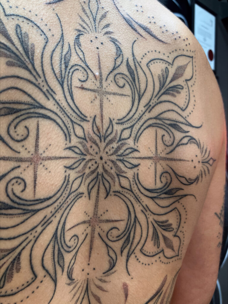 Close-up van een fijnmazige ornamentele tattoo op een schouderblad. Het ontwerp toont de precisie van de handpoked techniek met zachte dotwork-schaduwen en strakke, symmetrische lijnen in een ster-vormig patroon. Werk van Monica (Poked by Monique) op de Sint-Niklaas Tattoo Expo.
