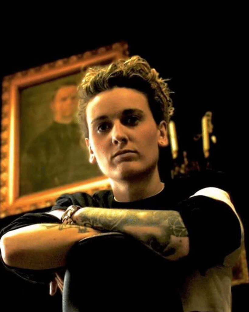 Tattoo-artiest Nikki Van Trappen poseert met gekruiste armen, waarbij haar eigen uitgebreide armtattoos zichtbaar zijn. Ze draagt een zwart-wit shirt en bevindt zich in een sfeervolle ruimte met een klassiek schilderij en een kandelaar op de achtergrond.