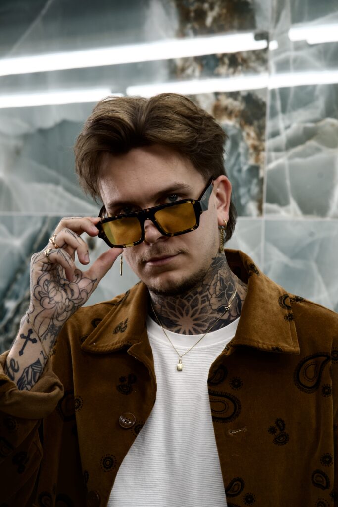 Een studio-opname van tattoo-artiest Kamil Jaster. Hij kijkt over de rand van zijn geel-getinte bril en draagt een bruine corduroy jas met paisley-motief. Zijn hals en handen zijn volledig bedekt met gedetailleerde tattoos, waaronder een groot mandala-patroon op zijn nek.