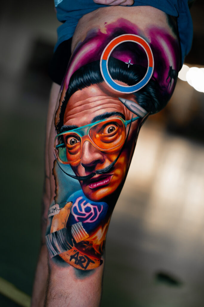 Een spectaculaire full color tattoo op een bovenbeen van Salvador Dalí in een moderne, surrealistische stijl. Dalí draagt een felblauwe en oranje bril, en de compositie bevat diverse abstracte elementen zoals een banaan met het woord ART en een zwevende cirkel. Werk van Kamil Jaster op de Sint-Niklaas Tattoo Expo.