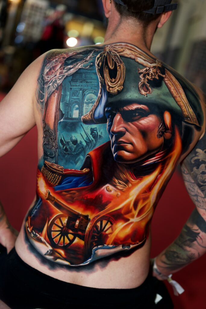 Een indrukwekkend, grootschalig rugstuk dat Napoleon Bonaparte afbeeldt in volle strijd. Het ontwerp barst van de felle oranje en rode vlammen rondom een kanon, met op de achtergrond de Arc de Triomphe en silhouetten van marcherende soldaten. Vakmanschap van Kamil Jaster tijdens de Sint-Niklaas Tattoo Expo.