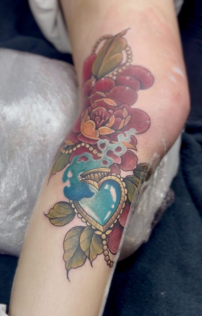Een opvallende neo-traditional tattoo op een onderarm. Het toont een menselijke schedel omringd door felle rode bloemen en groene bladeren. Het ontwerp maakt gebruik van sterke contrasten en een levendig kleurenpalet. Gezet door Beertje Malou op de Sint-Niklaas Tattoo Expo.