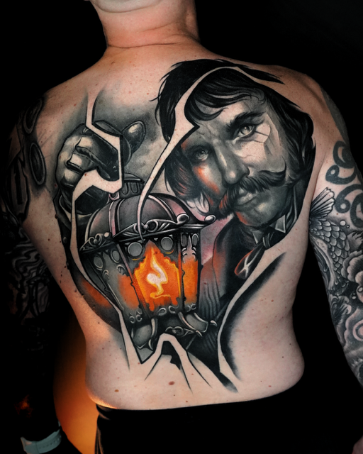 Een indrukwekkend rugstuk in een realistische stijl. De tattoo toont een gedetailleerde lantaarn met een gloeiende oranje vlam, geflankeerd door een portret van een man met een markante snor. De compositie maakt gebruik van een 'gebroken glas' effect en diepe contrasten. Werk van Tom van Oevelen op de Sint-Niklaas Tattoo Expo.