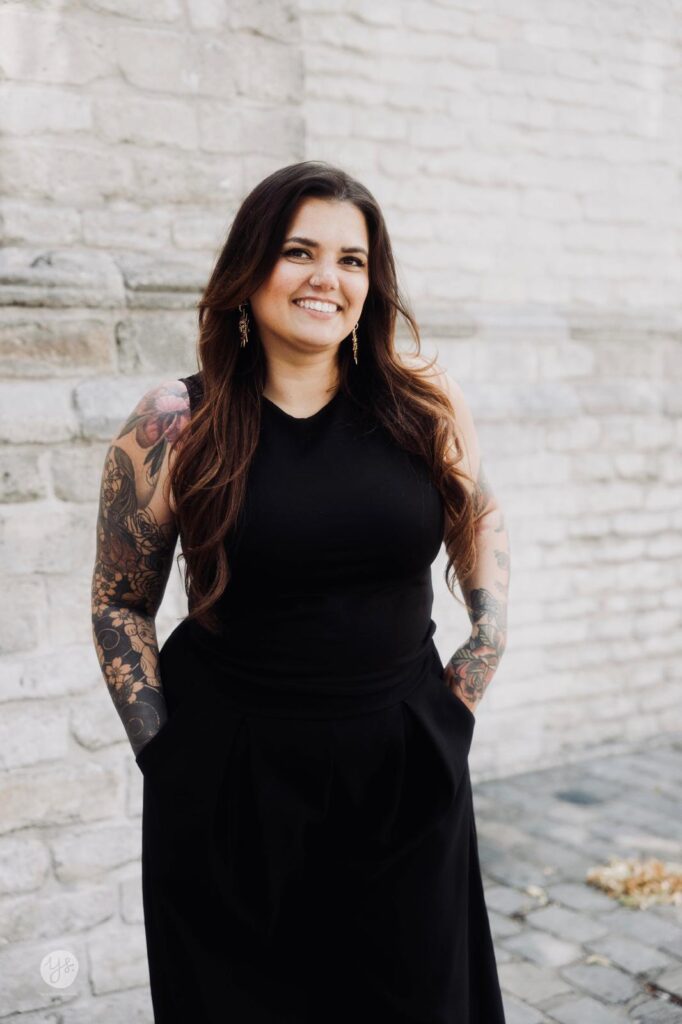 Een sfeervolle actiefoto van tattoo-artiest Beertje Malou op de Sint-Niklaas Tattoo Expo. Ze is geconcentreerd bezig met het tatoeëren van de arm van een klant. Beertje Malou heeft lang donker haar en draagt zwarte handschoenen en een zwart t-shirt. Haar eigen uitgebreide armtattoos zijn goed zichtbaar.