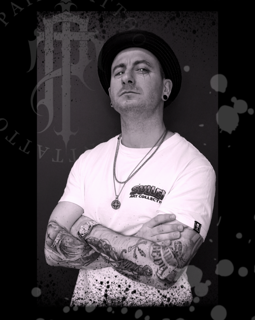 Een artistieke zwart-wit foto van tattoo-artiest Tom van Oevelen. Hij draagt een witte T-shirt, een donker hoedje en zilveren kettingen met een kompas-hanger. Hij poseert met gekruiste armen, waardoor zijn uitgebreide armtattoos goed zichtbaar zijn. Op de achtergrond is het logo van Tpain Tattoo subtiel verwerkt.