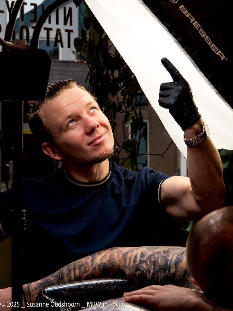 Een actiefoto van tattoo-artiest Ivan Gordienko tijdens de Sint-Niklaas Tattoo Expo. Hij kijkt met een glimlach omhoog naar zijn professionele Bresser softbox-verlichting terwijl hij een klant tatoeëert. Ivan draagt zwarte handschoenen en een horloge; op de voorgrond is een arm met een grote black and grey tattoo zichtbaar.